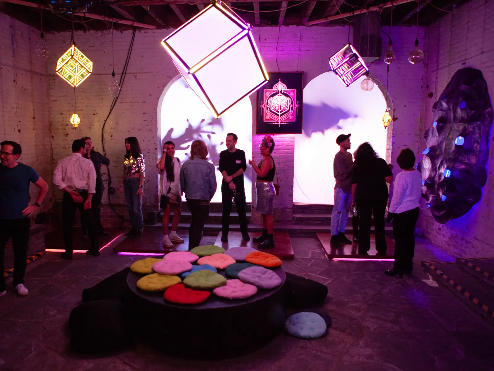 A circular haptic platform laid with coloured pads sits at the centre of a warehouse room, illuminated cubes hanging above and guests standing at the edges.